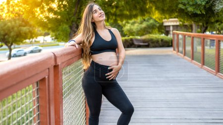 Foto de Retrato de una mujer embarazada sonriente en ropa deportiva apoyada en la valla, tocándose el vientre y mirando hacia arriba en el puente a pie al aire libre. - Imagen libre de derechos