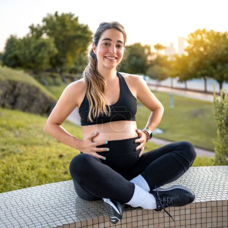Téléchargez les photos : Belle jeune femme enceinte en vêtements de sport toucher son ventre et assis à l'extérieur au coucher du soleil - en image libre de droit