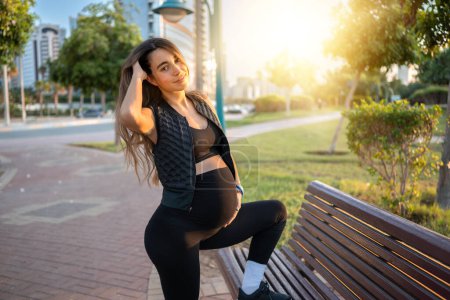 Téléchargez les photos : Belle jeune maman enceinte posant avec la jambe sur un banc dans le parc au coucher du soleil - en image libre de droit
