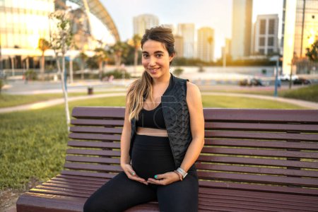 Photo for Portrait of friendly young future mother holding and touching her stomach while resting on bench outdoors - Royalty Free Image