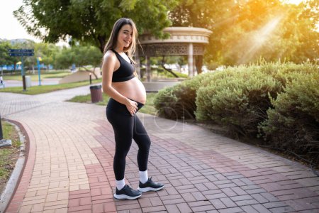 Foto de Retrato de larga duración de una mujer embarazada de nueve meses en ropa deportiva mostrando su gran vientre al aire libre al atardecer - Imagen libre de derechos