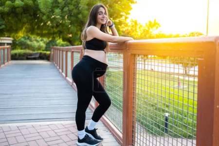 Téléchargez les photos : Portrait complet d'une femme enceinte souriante en tenue de sport appuyée sur une clôture et regardant la caméra. Sporty future maman dans le parc au coucher du soleil. - en image libre de droit