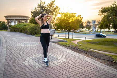 Photo for Full length portrait of young pregnant woman in sportswear tying hair and preparing for outdoor jogging and relaxing exercises in the park. - Royalty Free Image