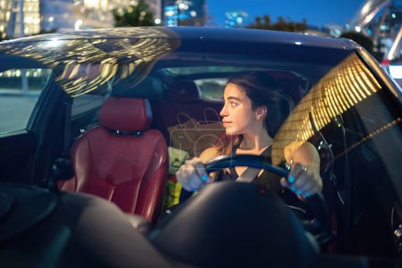 Photo for Front view of young beautiful woman driving car with shopping bags in back sits at night. Shopping, modern lifestyle, work and transportation concept. - Royalty Free Image