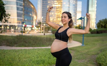 Téléchargez les photos : Portrait de femme enceinte sportive en tenue de sport montrant les muscles, debout en plein air dans le parc le soir. Mode de vie sain, sport et concept de grossesse. - en image libre de droit