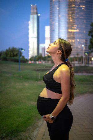 Téléchargez les photos : Vue latérale du profil d'une femme enceinte de neuf mois en tenue de sport noire regardant dans la rue de la ville - en image libre de droit