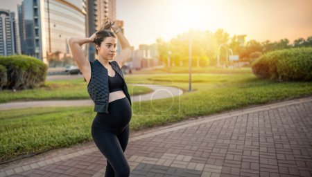 Téléchargez les photos : Jeune femme enceinte attacher les cheveux tout en ayant détendu marcher dans le parc au coucher du soleil. - en image libre de droit