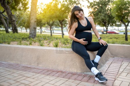 Téléchargez les photos : Belle femme enceinte assise sur un mur de pierre et relaxante dans le parc au coucher du soleil - en image libre de droit