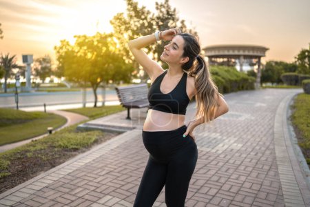 Téléchargez les photos : Belle jeune femme enceinte en tenue de sport debout dans le parc au coucher du soleil - en image libre de droit