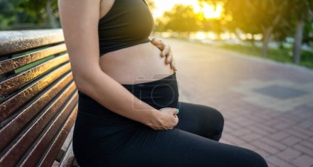 Téléchargez les photos : Gros plan du ventre de la femme enceinte assis sur un banc en bois à l'extérieur pendant le coucher du soleil - en image libre de droit