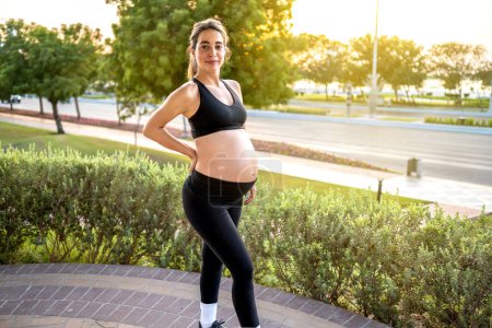 Téléchargez les photos : Charmante femme enceinte en tenue de sport debout à l'extérieur. Grossesse, accouchement, mode de vie sain - en image libre de droit