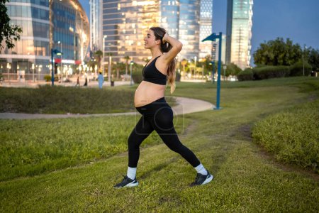 Foto de Vista lateral de la mujer embarazada en ropa deportiva haciendo saltos en un parque por la noche - Imagen libre de derechos