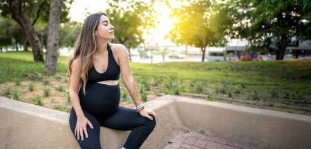 Foto de Hermosa mujer embarazada sentada en la pared de piedra y relajándose en el parque al atardecer - Imagen libre de derechos