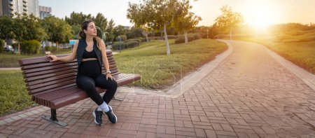 Téléchargez les photos : Jeune femme enceinte reposant sur un banc de parc en bois au coucher du soleil. Espace de copie - en image libre de droit