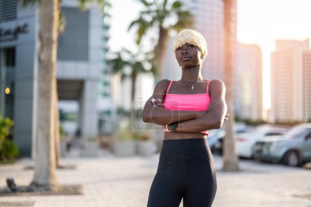Photo for Portrait of beautiful confident blond haired young black woman in sportswear with arms crossed posing on city street at sunset. - Royalty Free Image