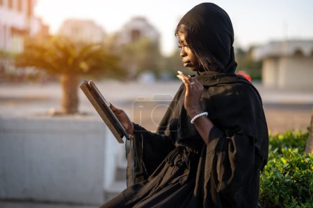 Foto de Retrato de una hermosa joven africana en ropa Abaya usando tableta al aire libre y saludando con la mano su pantalla durante la videollamada. - Imagen libre de derechos