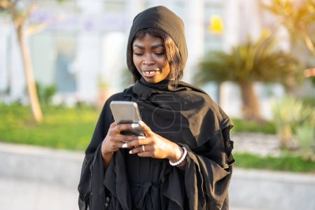 Foto de Retrato de una hermosa mujer africana usando abaya usando teléfono inteligente al aire libre - Imagen libre de derechos