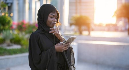 Photo for Portrait of beautiful African woman wearing abaya using smart phone outdoors - Royalty Free Image