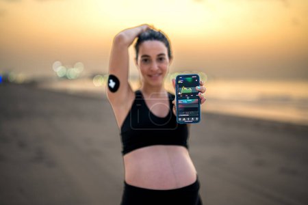 Photo for Beautiful pregnant woman using Continuous Glucose Monitor to improve diabetes control. Sporty pregnant woman showing phone displaying blood sugar test on smartphone App. - Royalty Free Image