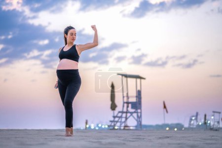 Foto de Mujer embarazada de nueve meses en ropa deportiva mostrando músculos, de pie en la playa de arena al atardecer. Estilo de vida saludable, deporte y concepto de embarazo. - Imagen libre de derechos