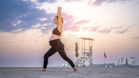Téléchargez les photos : Portrait sur toute la longueur de la femme enceinte en tenue de sport noire faisant pose de yoga Crescent Lunge sur la plage au coucher du soleil. Entraînement, yoga et concept de grossesse. - en image libre de droit