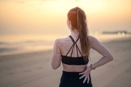 Mujer atlética en ropa deportiva de pie a la orilla del mar frotando los músculos de su espalda baja