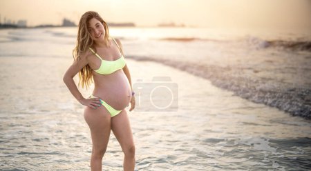 Téléchargez les photos : Belle femme enceinte en maillot de bain posant sur la plage au coucher du soleil - en image libre de droit