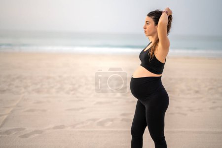 Téléchargez les photos : Jeune femme enceinte en tenue de sport attachant les cheveux et se préparant à une formation détendue sur la plage - en image libre de droit
