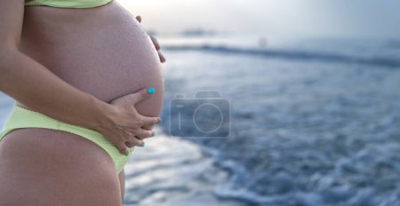 Téléchargez les photos : Gros plan sur le ventre d'une femme enceinte portant un bikini jaune sur la plage - en image libre de droit