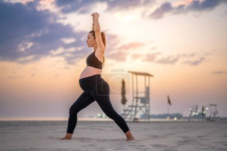 Téléchargez les photos : Vue latérale de la femme enceinte en tenue de sport noire faisant pose de yoga Crescent Lunge sur la plage au coucher du soleil. Entraînement, yoga et concept de grossesse. - en image libre de droit