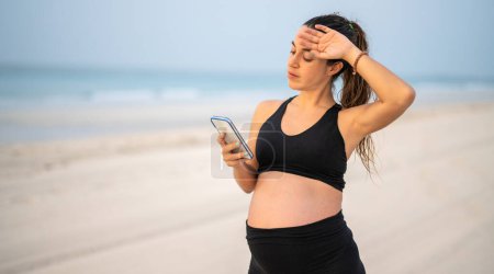 Photo for Beautiful pregnant woman standing over sandy beach seacoast looking at smart phone feeling tired holding hand on face - Royalty Free Image