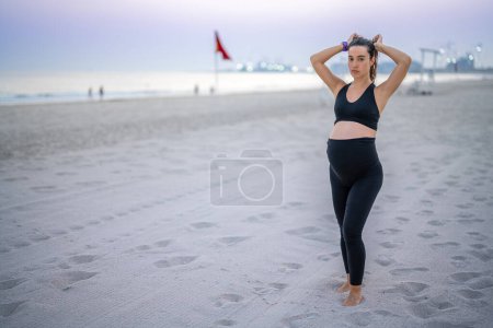 Téléchargez les photos : Jeune femme enceinte en tenue de sport attachant les cheveux et se préparant à une formation détendue sur la plage - en image libre de droit