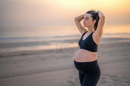 Téléchargez les photos : Portrait de jeune femme enceinte en tenue de sport attachant les cheveux et se préparant à un entraînement détendu sur la plage au coucher du soleil - en image libre de droit