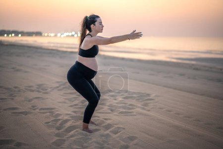 Foto de Vista completa de una joven embarazada haciendo ejercicio y haciendo sentadillas en la playa de arena al atardecer - Imagen libre de derechos