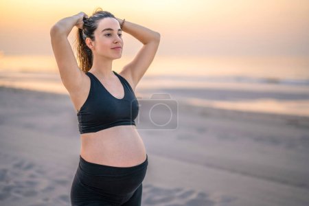 Photo for Portrait of young pregnant woman in sportswear tying hair and preparing for relaxed training on the beach at sunset - Royalty Free Image