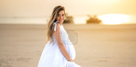 Photo for Profile view of pregnant woman with long hair in white dress on the beach at sunset - Royalty Free Image
