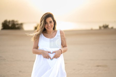 Photo for Pregnant woman holding tummy and showing heart shape with hands at beach - Royalty Free Image