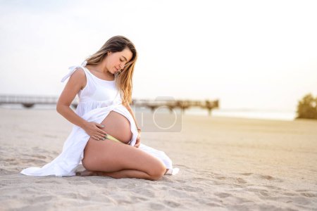 Pregnant woman wearing white dress sitting over sea shore outdoors. Motherhood. Maternity. Healthy lifestyle