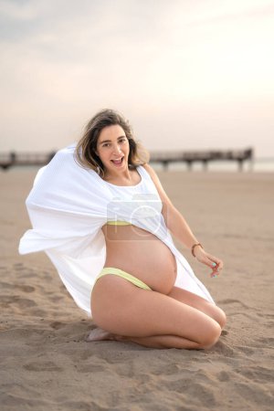 Foto de Foto vertical de la mujer embarazada salida en vestido blanco sentado en la playa - Imagen libre de derechos