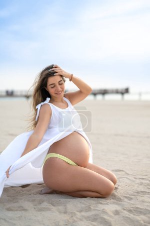 Téléchargez les photos : Portrait complet d'une jeune femme enceinte assise jouissant d'un vent estival sur la plage. Concept de grossesse, de maternité et d'attente du bébé. - en image libre de droit