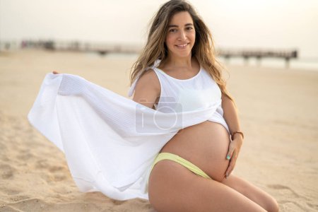 Téléchargez les photos : Portrait de femme enceinte en robe blanche assise sur le sable à la plage - en image libre de droit
