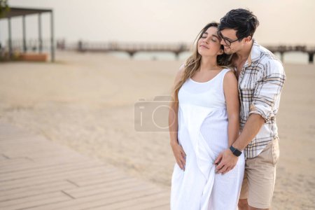 Foto de Hombre guapo abrazando a su esposa embarazada en vestido blanco en la playa durante el atardecer - Imagen libre de derechos