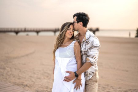 Photo for Happy embraced couple at beach. Handsome man hugging his pregnant wife and enjoying together tranquil moments at sunset - Royalty Free Image