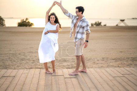 Schöne schwangere Frau in weißem Kleid tanzt mit ihrem hübschen Mann am Strand. Junges Paar hat Spaß im Freien