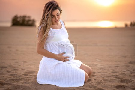 Téléchargez les photos : Belle future maman assise sur le sable de la plage et touchant son ventre au coucher du soleil - en image libre de droit
