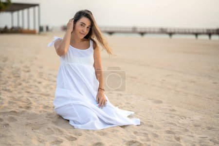 Photo for Young pregnant woman in white dress sitting on sand at beach - Royalty Free Image