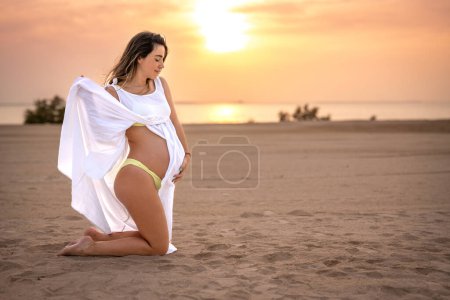 Téléchargez les photos : Vue latérale de la belle femme enceinte en robe blanche debout sur les genoux, tenant son estomac et profitant de la côte de la plage océanique au coucher du soleil. - en image libre de droit