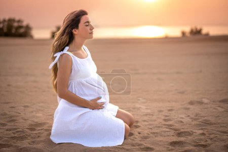 Téléchargez les photos : Femme enceinte réfléchie et détendue assise sur le sable et méditant sur la plage au coucher du soleil - en image libre de droit