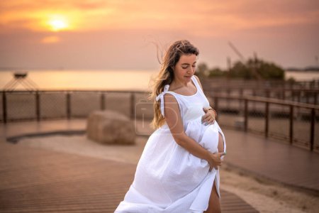 Téléchargez les photos : 9 mois femme enceinte portant une robe blanche et touchant son ventre sur le trottoir de la plage au coucher du soleil - en image libre de droit