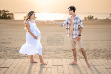 Téléchargez les photos : Amour de la femme enceinte et bel homme en chemise déboutonnée atteignant les autres mains à la plage - en image libre de droit